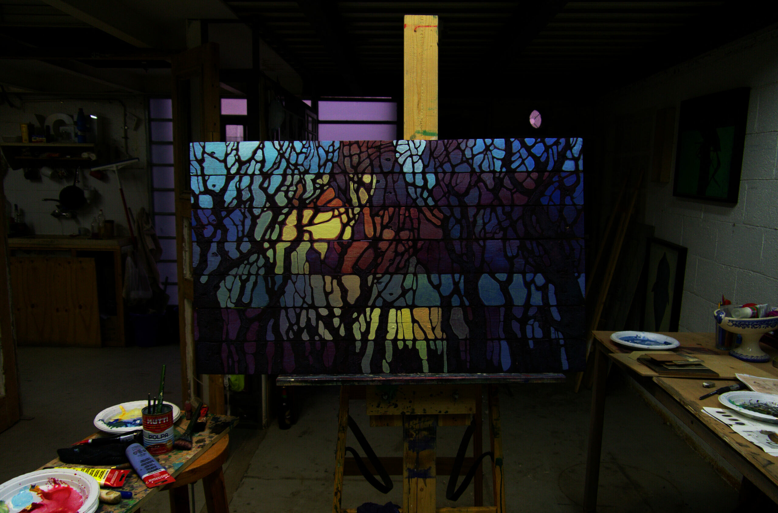 Painting showing the canvas 'The Lake' lit on a wooden easel in the dim studio, amid tables of paint, palettes and brushes - the score-painting in the place of its making.